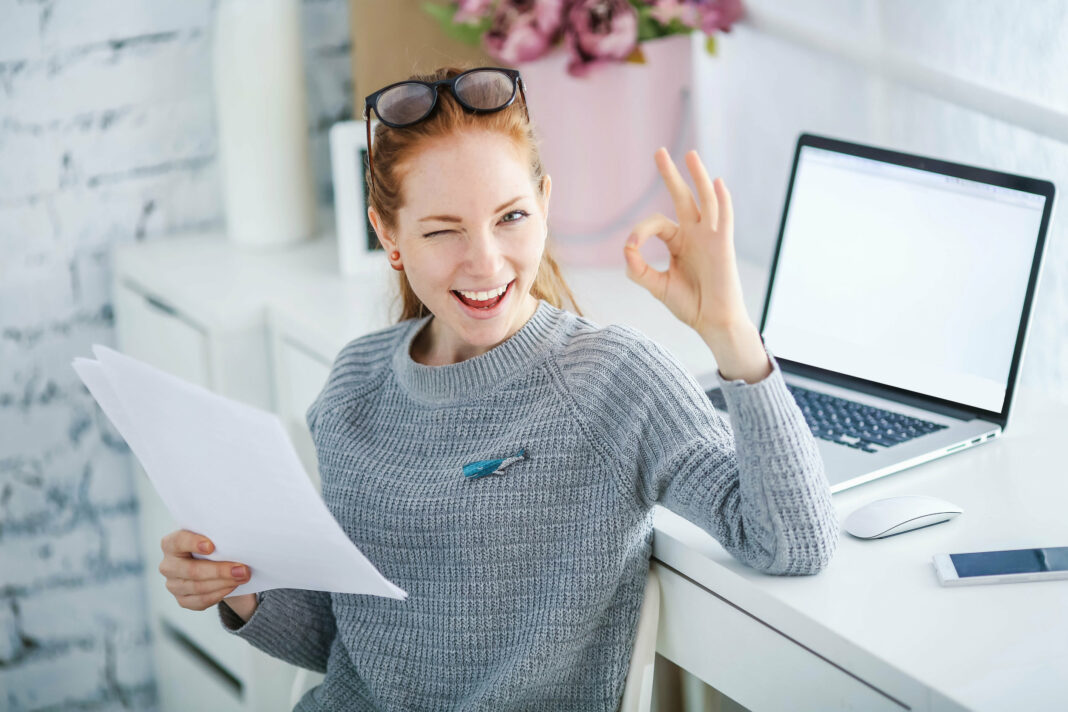 Young beautiful woman with red hair, wearing glasses, working in the office, uses a laptop and mobile phone