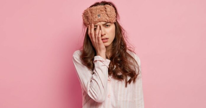 egy nő szemmaszkkal a fején fogja a fejét pizsamában