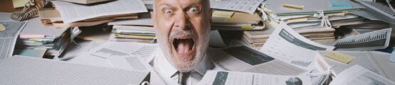 idősebb férfi betemetve számlákkal, papírokkal, száját nagyra nyitva ordít