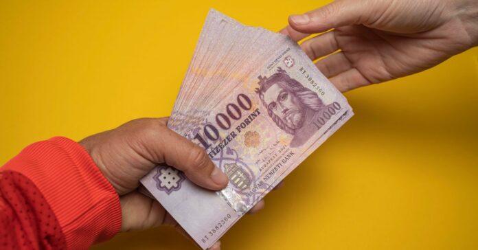 egy köteg tízezer forintos bankót nyújt ét egy kéz egy másiknak sárga háttér előtt