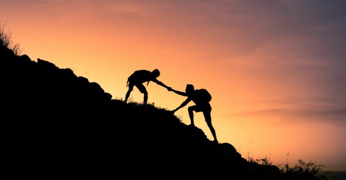 Two,Male,Hikers,Helping,Each,Other,Climb,Up,A,Mountain. Hóvége - Támogatáskereső