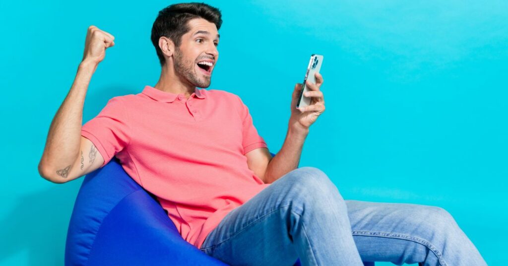 rózsaszínpólós férfi ül egy kék babzsákfotelben, mobilt tart bal kezében, jobbját felemeli, örül