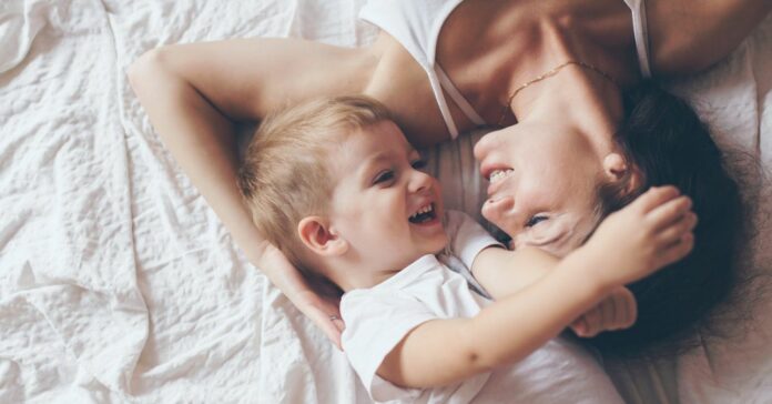 Fiatal, barna hajú anyuka 2 év körüli kisfiával fehér ruhában, fehér lepedőn fekve
