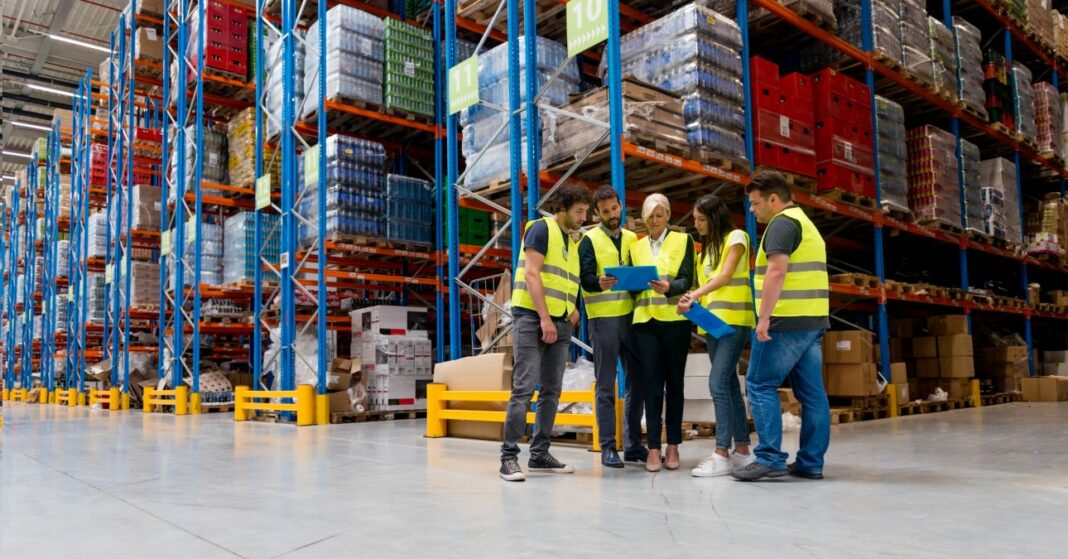 Egy raktárban munkások állnak az áruval megpakolt polcok előtt