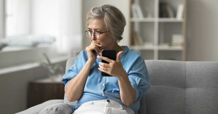 Idősebb, visszafogottan öltözött hölgy ül a szürke kanapén aggódva fogva mobiletelefonját
