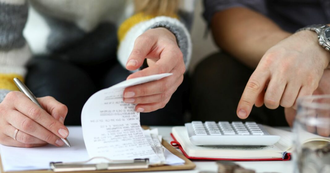 Közeli kép, az asztalon számológép, egy nő a blokkok között keres, egy férfi számokat üt be a számológépbe