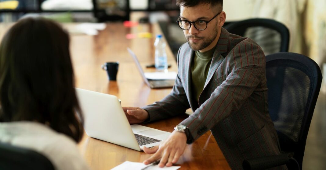 fiatal férfi íróasztalnál, egy papírt tol át egy nőnek