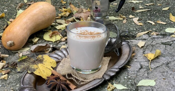 egy bögre pumpkin spice latte ezüst színű tálcán, mellette 2 vaníliarúd, őszi falevelek, a háttérben sütőtökkel