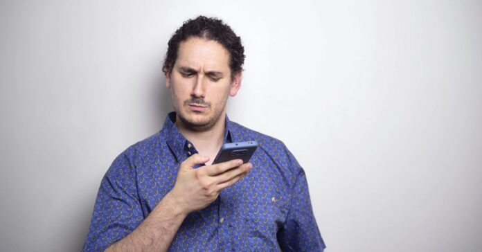 +39 hívószám csalás fiatal fehér férfi kék ingben gyanúsan néz a telefonjára