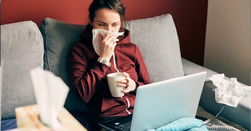 beteg nő az orrát fújja és teát iszik miközben laptopot tart az ölében