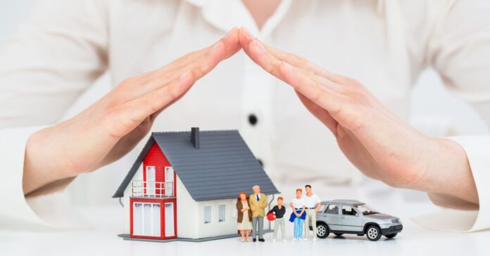 egy családi ház makettje családtagokkal és egy autóval, egy nő két kezét összetéve háztetőt formál felette