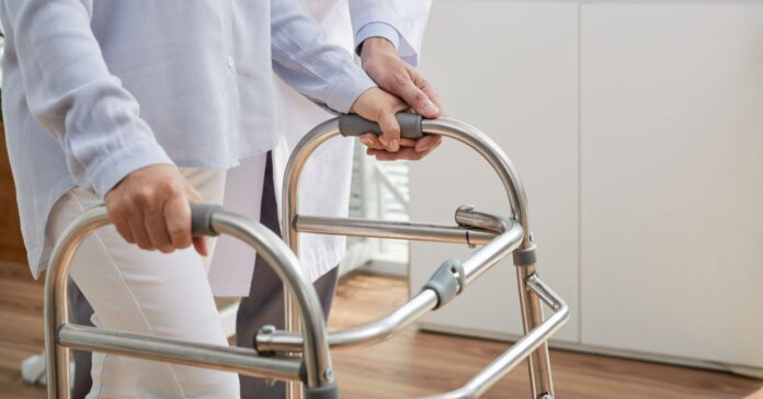 idős nő járókeretre támaszkodik, mellett egy fehér köpenyes kezelőorvosa nyújt segítséget