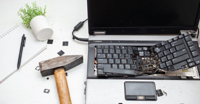 szétvert laptop az asztalon, mellet kalapács, felborult műnövény, toll és üreg jegyzettömb