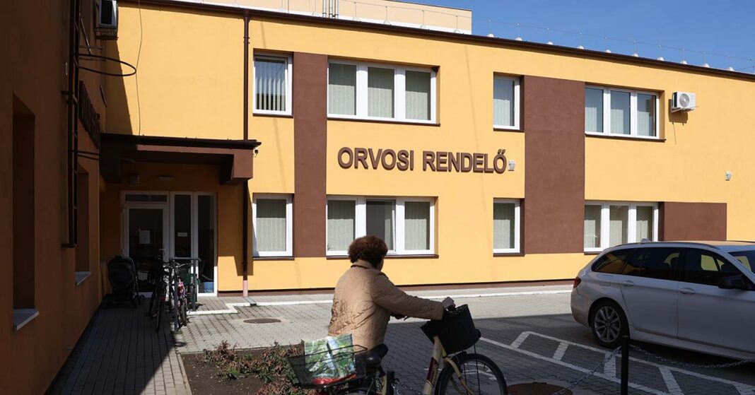 orvosi rendelő épülete, előtte biciklit tol egy idősebb nő