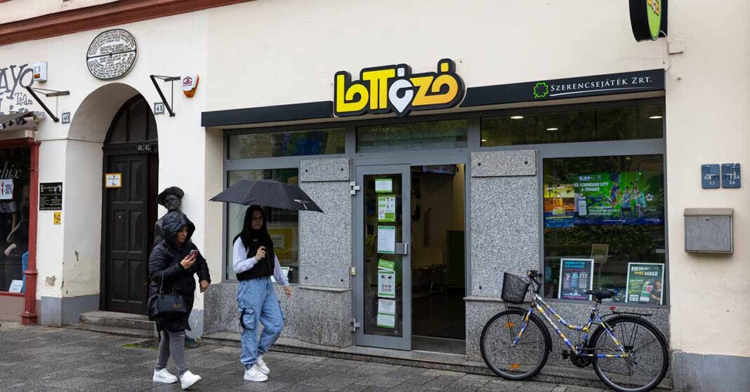 Lottózó bejárata előtt járókelők sétálnak el esernyővel