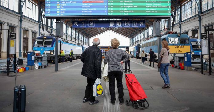 vasútállomáson kijelző, előtte két idős áll egy lidlös szatyorral és egy banyatankkal