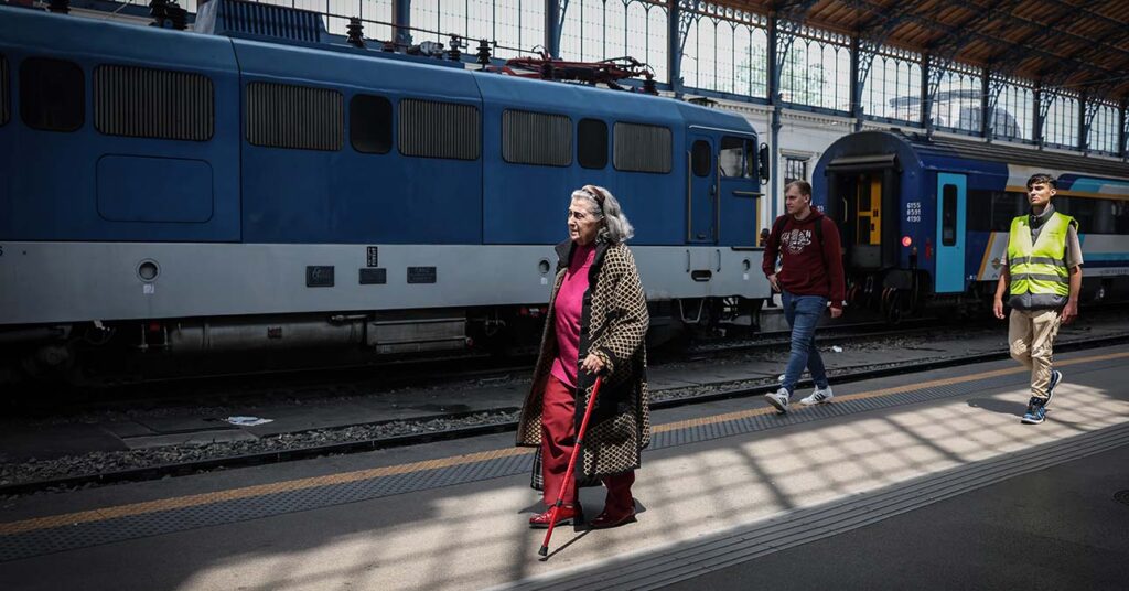 nyugdíjas néni fejkendőben, kabátban, bottal sétál a pályaudvaron