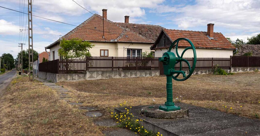 kistelepülés, kút, mögötte családi ház és üres telek