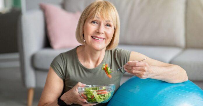 idősebb nő gimnasztikai labdára támaszkodik, közben egy tál salátát eszik mosolyogva
