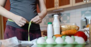 fiatal nő mérőszalaggal méri a derekát, előtte az asztalon élelmiszerek