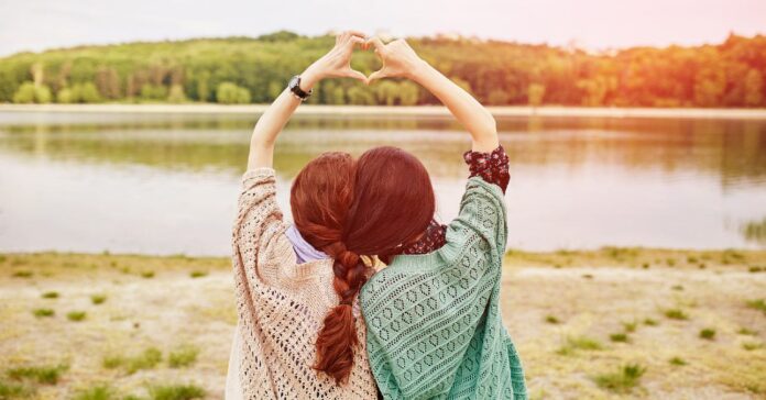 két nővér összevont hajjal, egymás mellett egy tó partján, kezükkel szívet formáznak