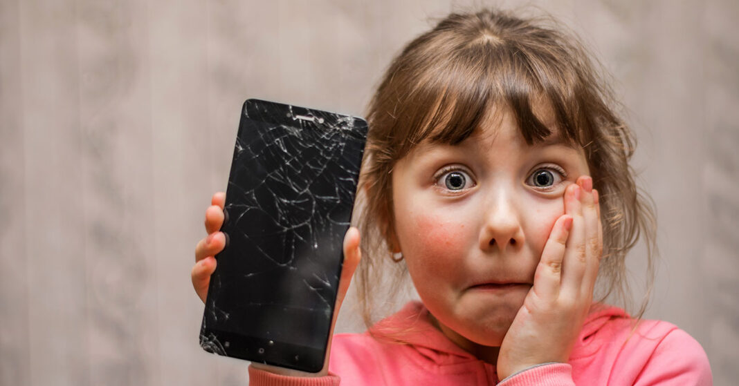 Gyermek törött mobiltelefont tart a kezében, bűnbánó arccal néz a kamerába