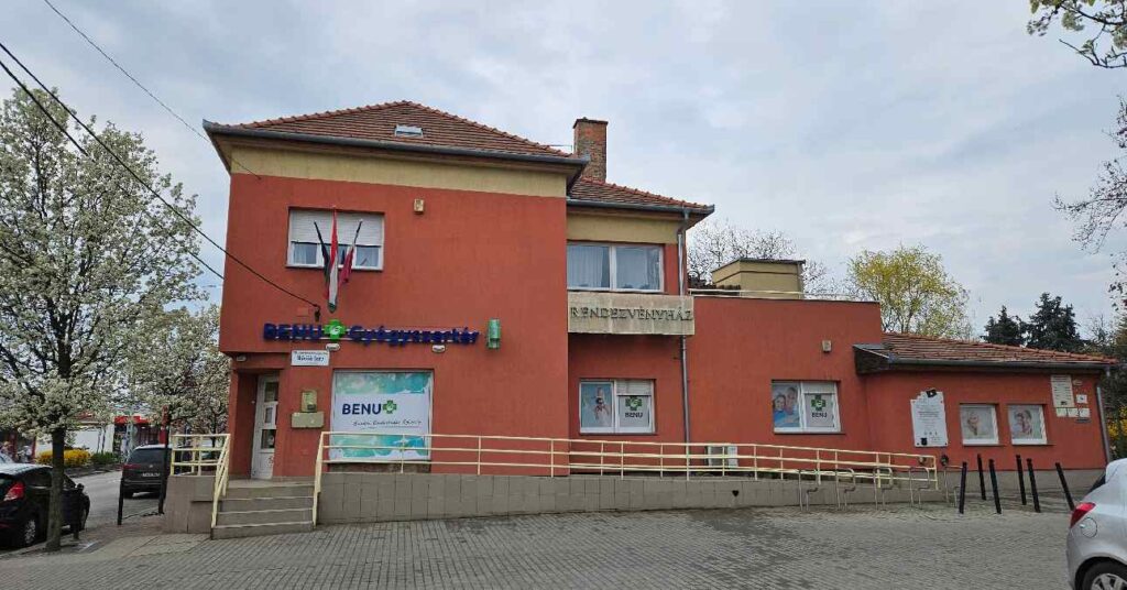 budapesti Benu gyógyszertár, kétszintes épület, téglaszínűre festve, előtte üres parkoló egyetlen autóval