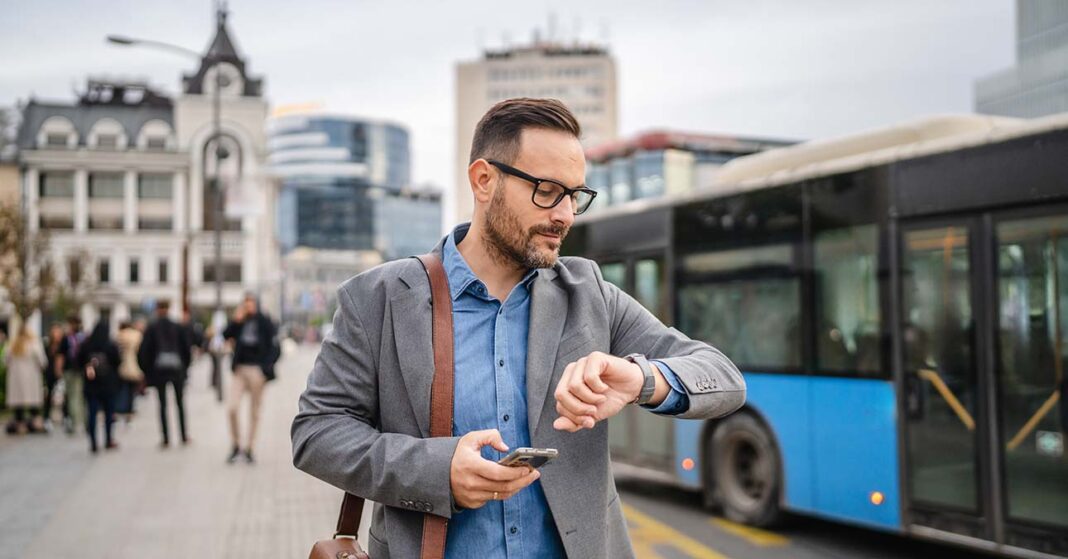 Középkorú férfi a telefonját használja az utcán, háttérben épületek és tömegközlekedés