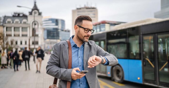 Középkorú férfi a telefonját használja az utcán, háttérben épületek és tömegközlekedés