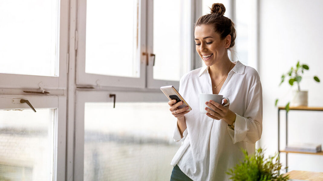 Vidám középkorú nő mobiltelefonnal és kávésbögrével áll egy nagy fehér ablak előtt