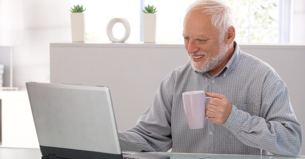Hide the Pain Herold, azaz nyugdíjas, ősz hajú férfi laptop előtt ül az íróasztalnál, bal kezében kávét fog