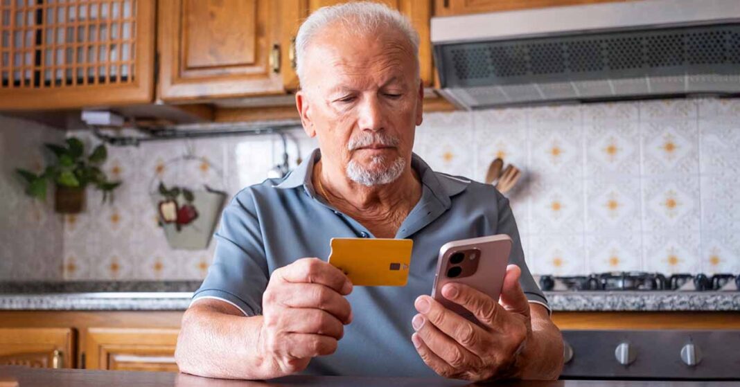 Nyugdíjas férfi webshopból rendel a konyhában ülve, kezében mobiltelefon és bankkártya